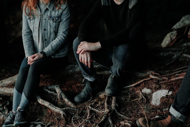 This is a picture of 3 people who are shown from the neck down. They are dressed in dark pants. The woman is in a denim jacket. They are in the woods sitting on a log.