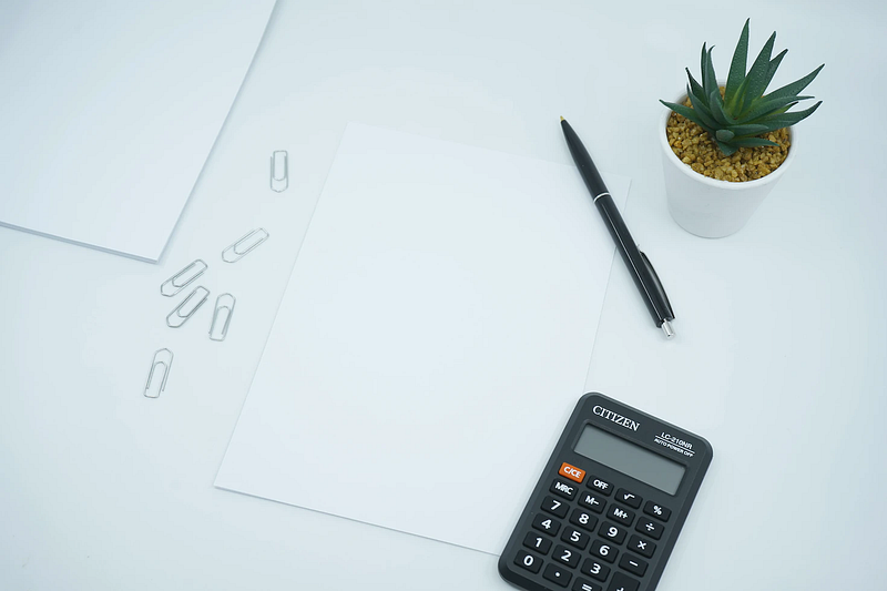 blank paper, pen, calculator, paper clips, and a potted succulent on a white table