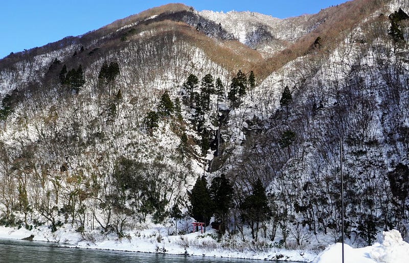 Stark red shrine gates at the bottom of a snowy hill along the Mogami River in Yamagata Prefecture
