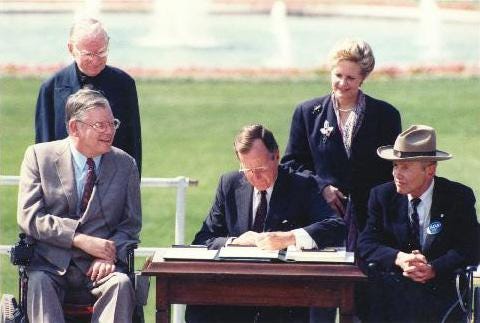 President Bush sits on a wooden desk and signs the ADA on the South Lawn (the lawn is green with a water fountain in the background). Behind him stands First Lady Barbara Bush. He is surrounded by Evan Kemp, Chairman of the Equal Employment Opportunity Commission, Justin Dart, Chairman of the President’s Committee on Employment of People with Disabilities; Rev. Harold Wilke, and Swift Parrino, Chairperson, National Council on Disability.