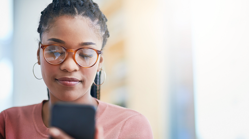 Woman using smartphone — A young woman with braided hair and glasses wearing a pink sweater looks at her smartphone with a focused expression. The background is blurred.