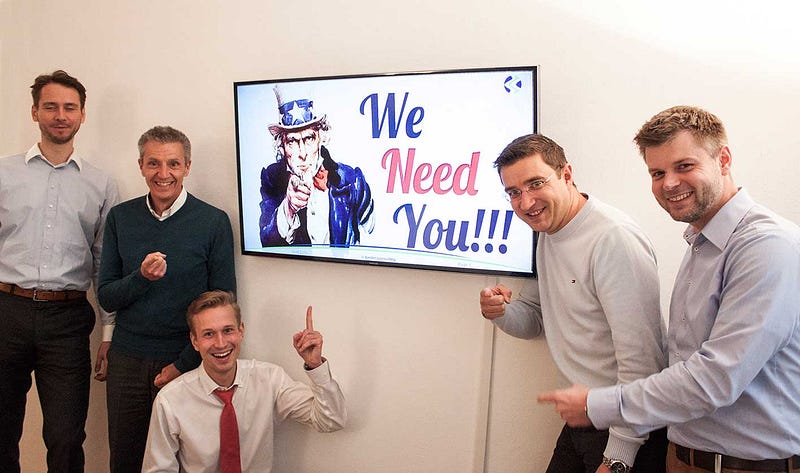Five men standing next to a TV with the iconic image "we need you"