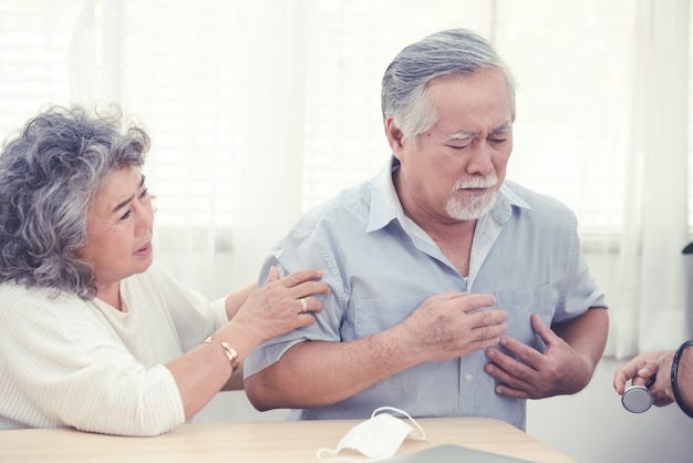 An elderly man clutching his chest in pain, showing signs of a possible heart attack, while an elderly woman beside him looks concerned and supports him. A doctor with a stethoscope is partially visible, approaching to provide medical assistance. The setting appears to be a home or clinic with natural light coming through the windows.