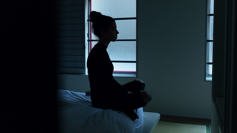 A young woman sits bolt upright in her bed at night. We see her from the side, a window in the background.