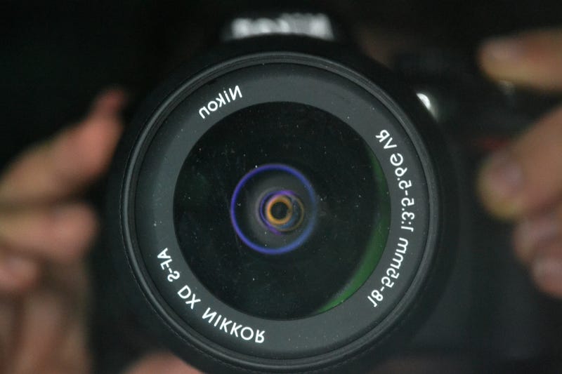 Fotografia frontal, plana fechado de mãos segurando uma câmera. Mãos desfocadas e lente da câmera focada.