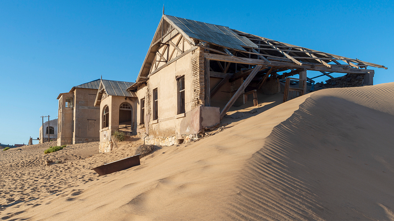 Abandoned buildings in the desert — A row of crumbling buildings with collapsed roofs partially buried in sand dunes, under a bright blue sky. The structures appear old and abandoned.