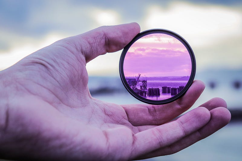 Reframing: a hand holding a purple-tinted lens showing an altered landscape beyond