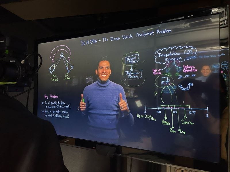 A man in a blue turtleneck gives a thumbs-up standing behind a Lightboard covered in mathematical equations.