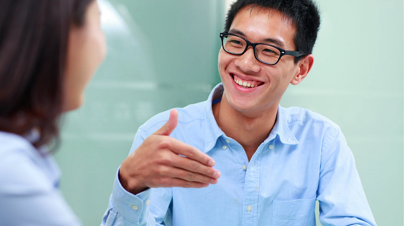 Smiling man in conversation — A young man wearing glasses and a light blue shirt smiles while engaging in conversation with another person, gesturing with his hand.