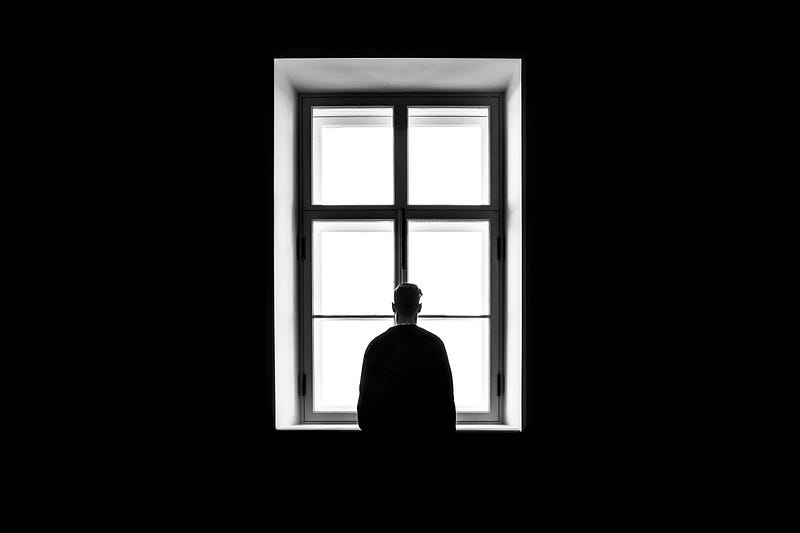 Black and white image (from behind) of a person in silhouette looking outside of a window.