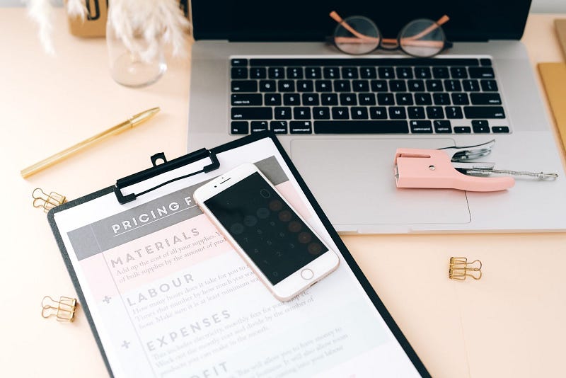 a pen, a laptop, a pair of round eyeglasses, a pink stapler, a clipboard, and a white smartphone on a peach colored desk