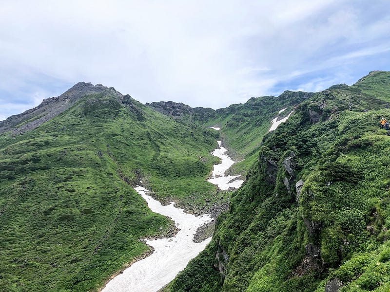 The green mountain valley with expansive snowbanks known as Senja-dani, the valley of 1,000 serpents, surrounded on both sides by rocky peaks, and the summit of Chokai-san (Mt. Chokai).
