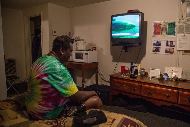 Jamerson sits on his bed in the one-room residence he rents at the Shamrock Motel on April 14, 2016. After a day spent on the street corner selling CDs, Jamerson watches the TV mounted on a wall he's decorated with memorabilia from his past. Photo by Kjell Redal 