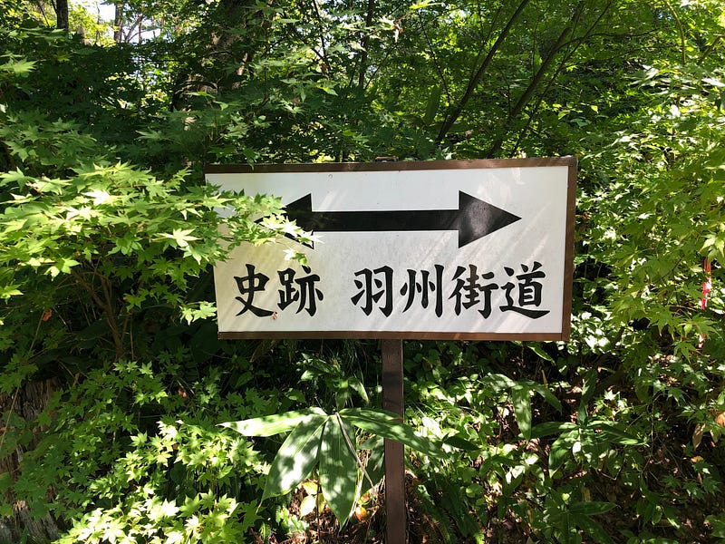 A white sign in the middle of a bunch of trees pointing the way of the historic Ushu Kaido.