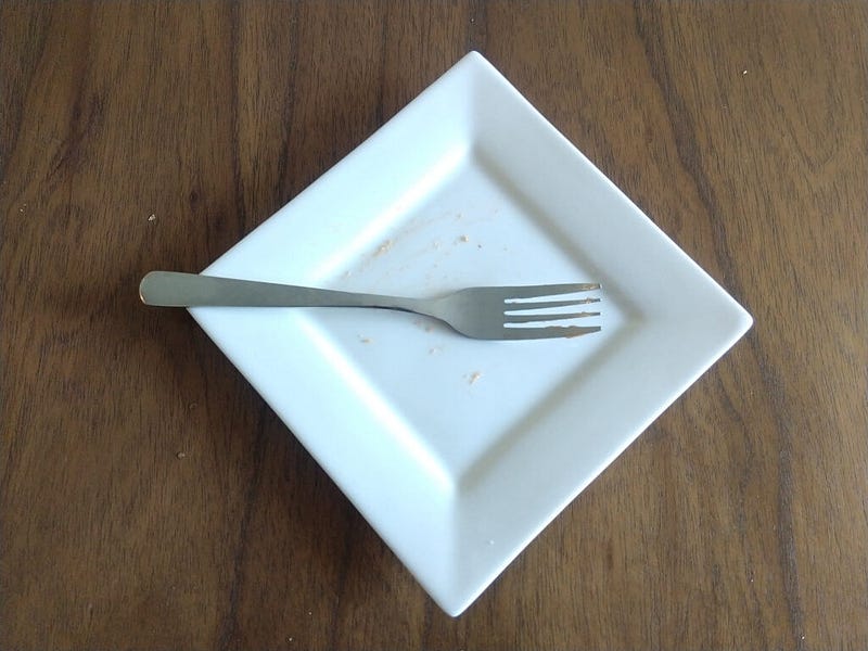An empty white plate, apart from a few crumbs, on a table with a fork resting on top of it.