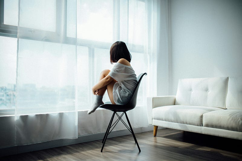 A young woman sits in a chair, her knees brought up to her chest, as she looks outside an apartment large window.