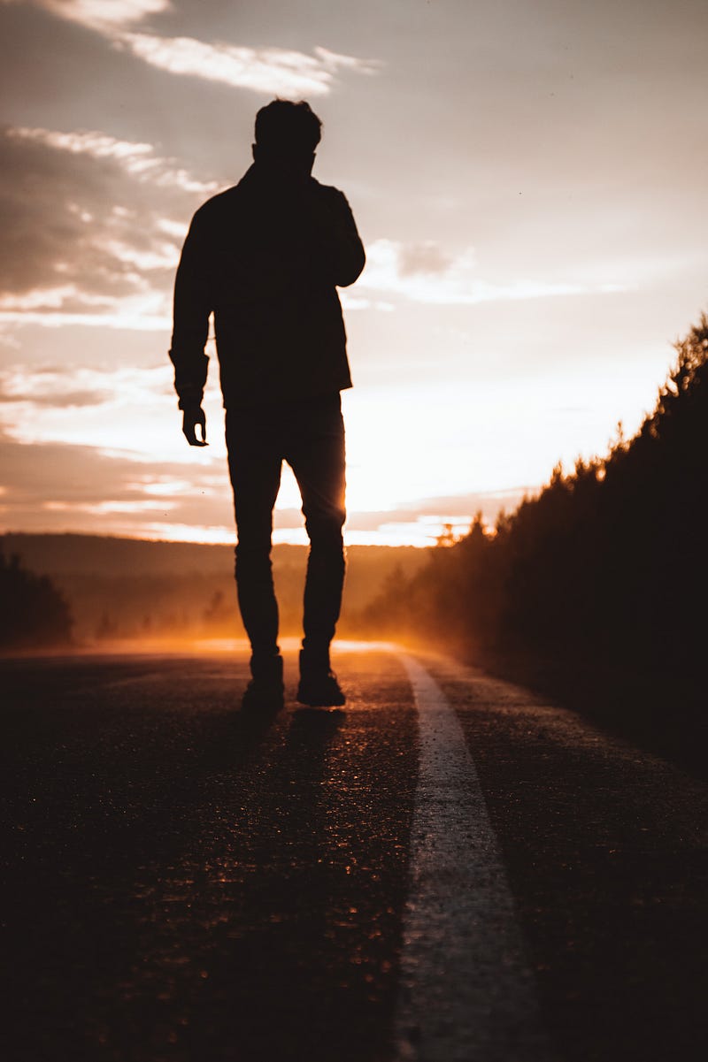 A man is on a deserted highway and he is walking towards the remaining light of the sun.