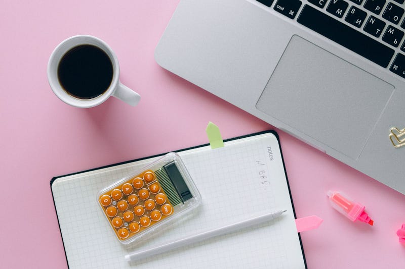 top view of a coffee mug, a laptop, a pink highlighter, a notebook. a calculator, and a pen, on top of a pink background