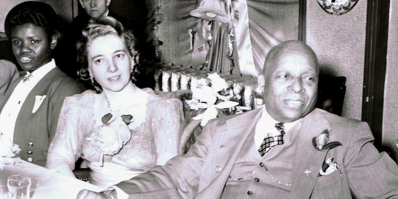 A black and white photo of a Black man in a suit sitting next to a white woman in a light lace dress at a banquet table. Both are smiling.