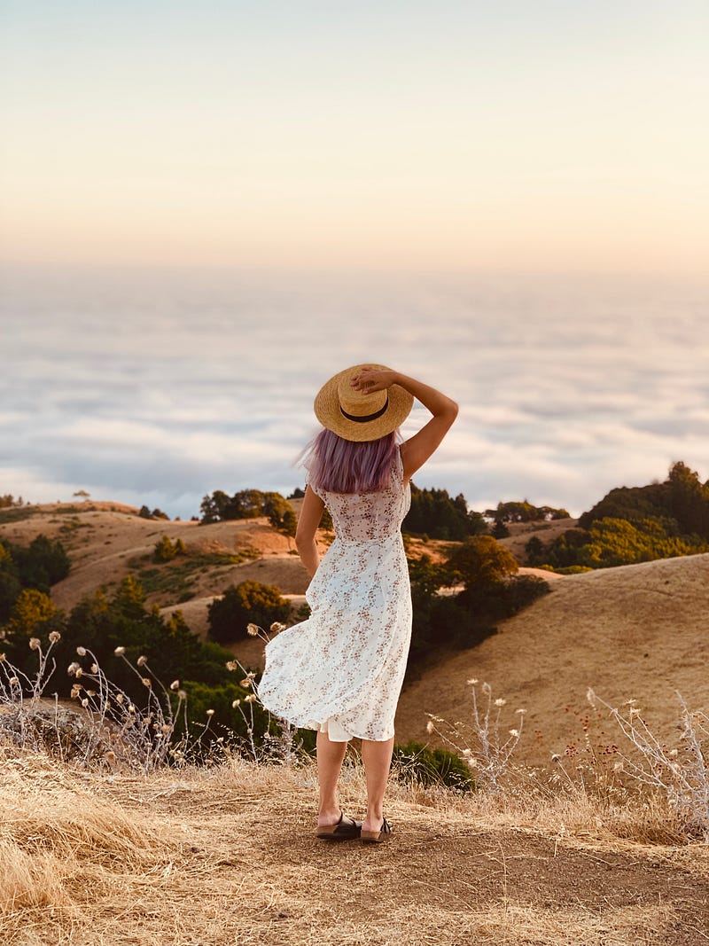 A lady wearing a white dress and a hat and looking to the beautiful clouds and the view.