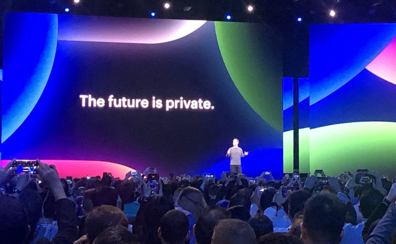 Mark Zuckerberg stands on stage in front of a large crowd, the words "The future is private" flashed on a screen behind him.