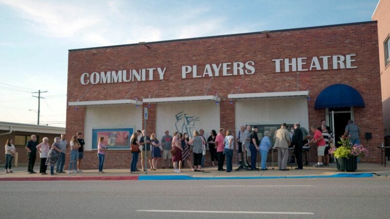 A long line of people waiting to see a show in front of a building with “Community Players Theatre” written across the top