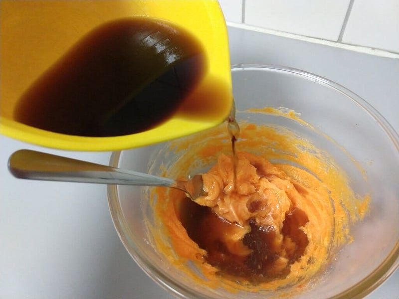 A yellow plastic bowl containing cola being tipped into a glass bowl over a mix of cream cheese and orange jelly crystals