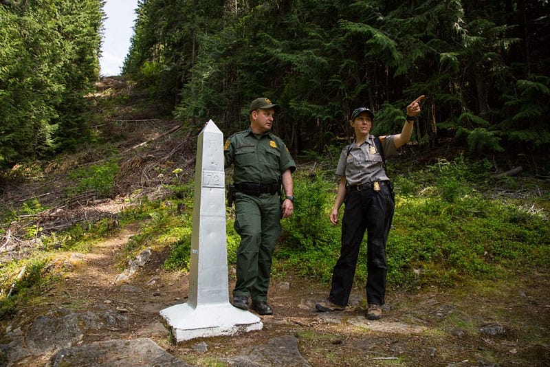 NPS ranger Jordan Mammel points border patrol agent Brooks Madden to a hillside where the clearing they stand on, demarcating the U.S.-Canada border, continues up the opposite side. The obelisk at Madden's side is number 72, with the first of them starting at the Pacific Ocean. 