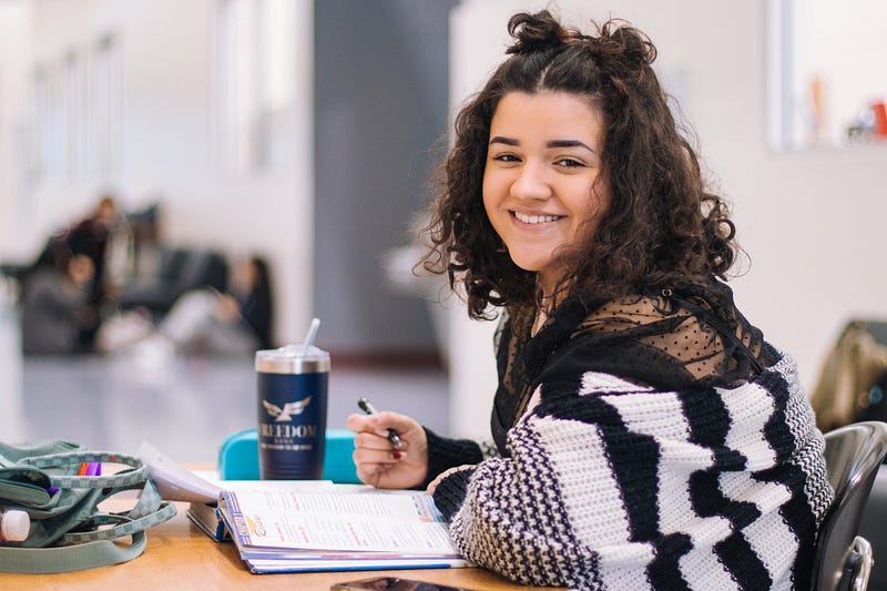Young teenage girl smiling at the camera. She’s holding a pen and writing in her notebook.