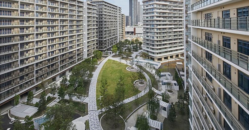 Image of the Tokyo Olympic Village. Tall, white buildings with glass windows surround a patch of a grassy area that includes tables, trees, and walk ways.