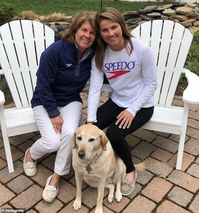 Becca and her mother/PCA, Maria Meyers smiling and sitting outdoors on white chairs with their heads together. Maria is wearing a blue pullover sweater, white pants, and off-white boat shoes. Becca is wearing a white long sleeve shirt with the SPEEDO logo, black leggings, and tan boat shoes. Becca’s guide dog sits in between them on the brick ground.