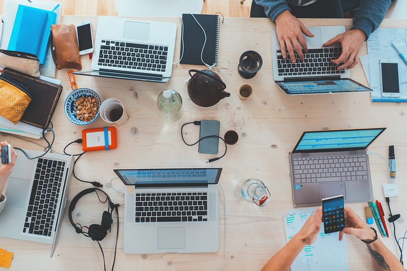 A table with multiple computers on it and people working on them.