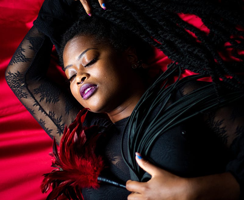 A womxn lies with her eyes closed on a red, silk blanket. Her lips are parted, and stained a metallic purple. Her dark dreadlocks lay around her head and shoulder. On her chest, and grasped in her hand, lies a cat o'nine whip.