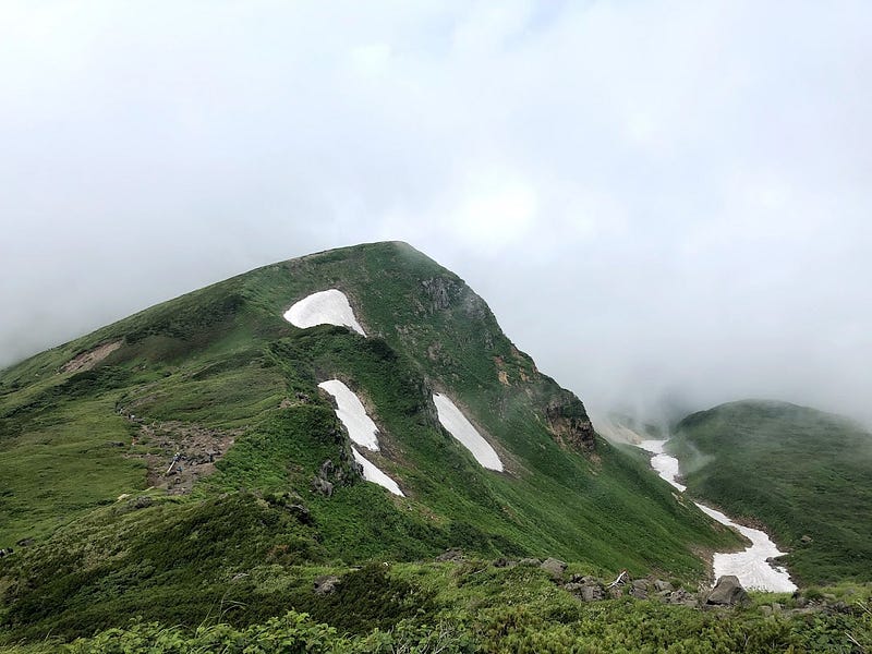 A spiky deep green peak covered in snowy patches protrudes through a cloudy sky on Chokai-san (Mt. Chokai)