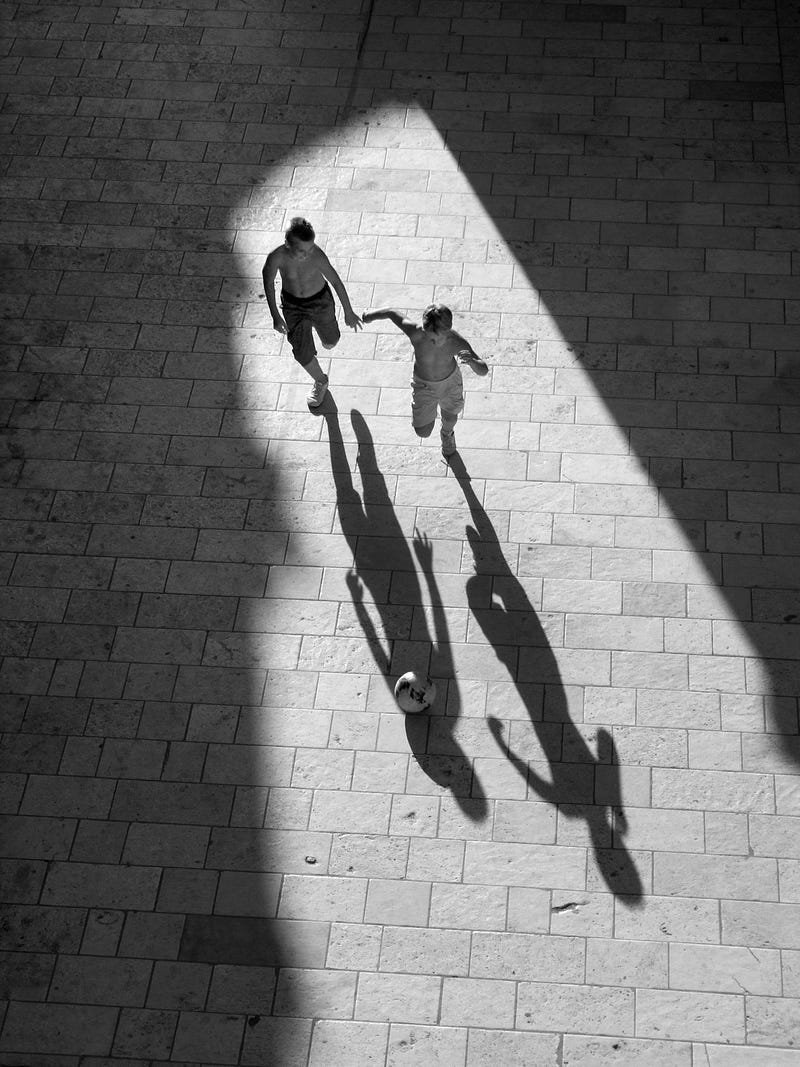 In a black and white image, two children approach, building shadows to the left and right of them. We see the kids from above as they hold hands and run towards us.