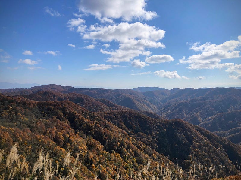 The view from the Akaha lookout on the autumn wonderland of Mount Taizo: blue sky dotted with clouds and mountains covered in the reds, yellows, and greens.