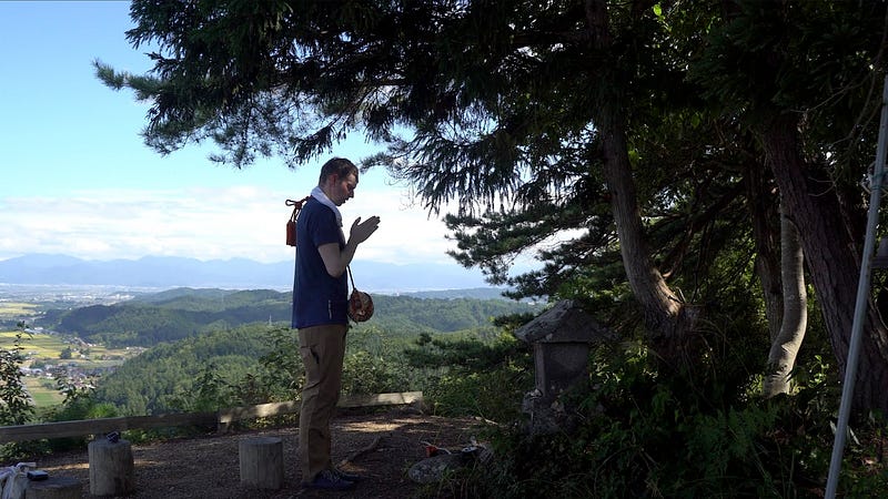 Tim Bunting AKA Kiwi Yamabushi praying to the shrine at the summit of Tengu-yama, mountain forests and rolling hills in the distance.