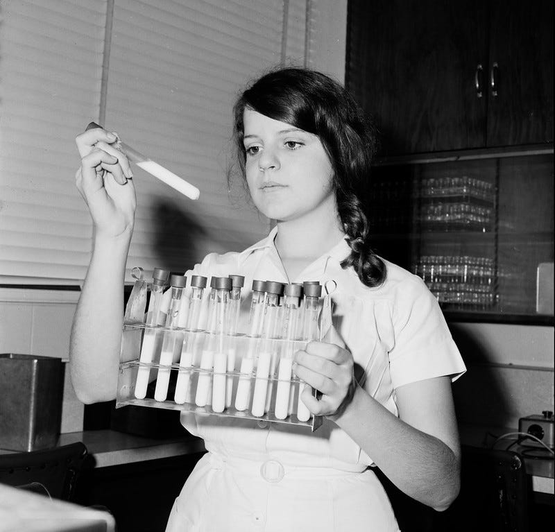 A photo of woman carrying glass milk sample phials. She’s holding one of the phials in front of her and looking at it.