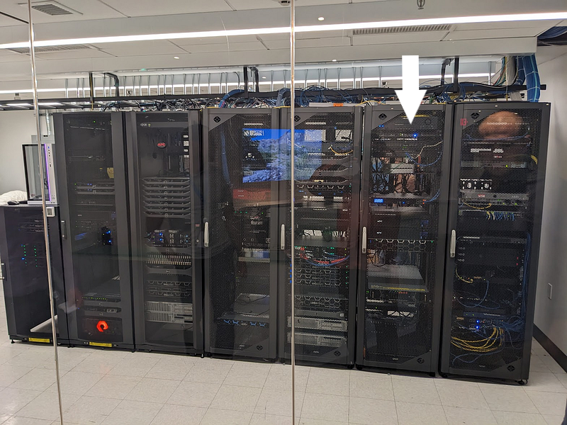 A row of large black server racks in a data center or server room. The racks are filled with various networking equipment, servers, and cables. Blue LED lights can be seen inside some of the racks. The room has a white tiled floor and ceiling with fluorescent lighting. There is an arrow pointing to the Quansight server.