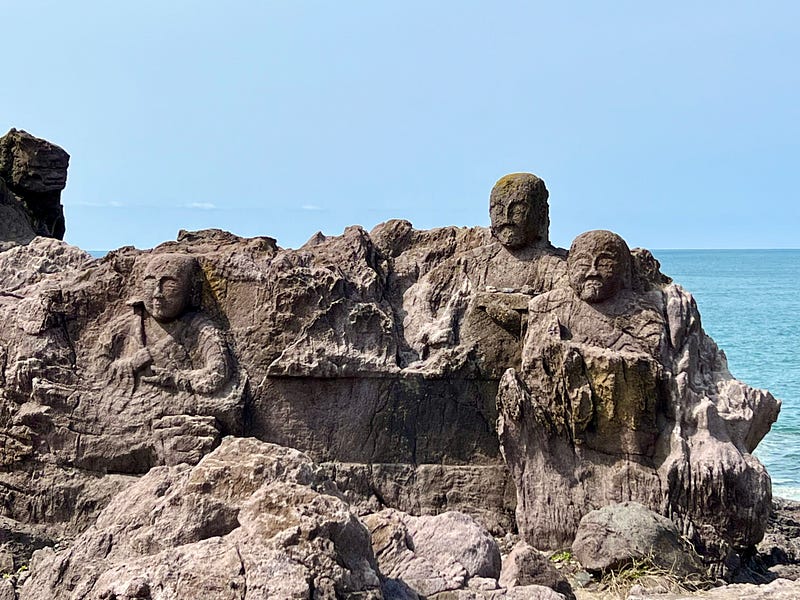 Three figures carved from volanic rock with the sea just behind them.