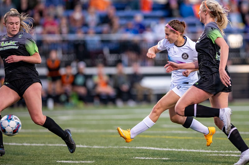 Soccer player takes shot between the legs of another, with a third in shot