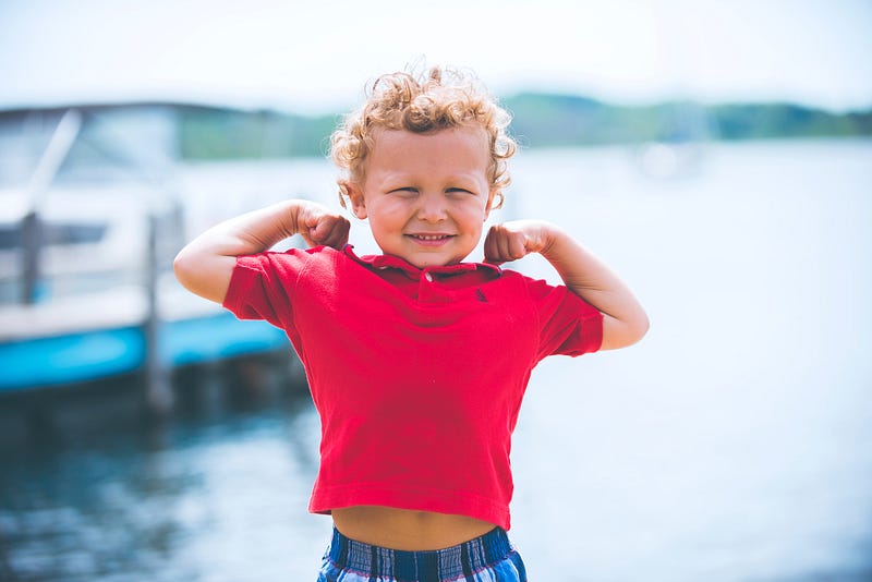 Toddler with curly blond hair wearing a red polo and flexing his arms. Water in the background.
