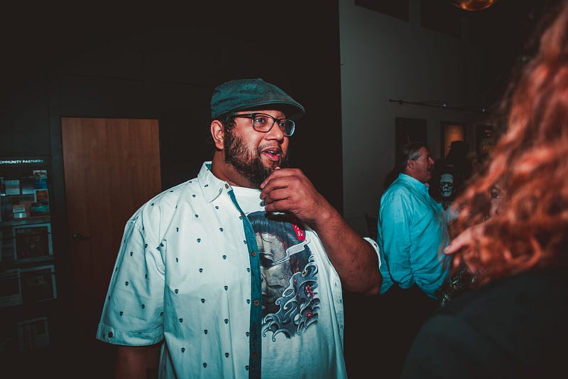 A black man in a cap, glasses and t-shirt with horror art rubs his beard while socializing in a dim, people-filled space.