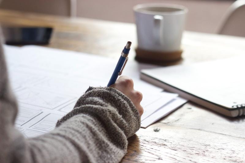 A photograph of a hand writing something at a desk, with papers strewn, a laptop closed next to them, and a coffee cup at the end of the desk.