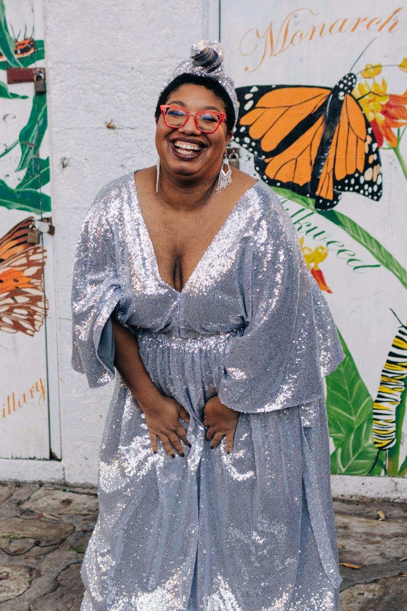 Sondra — a Black woman — poses in a sparkling silver gown. Behind her is a mural showing butterflies in bright colors.