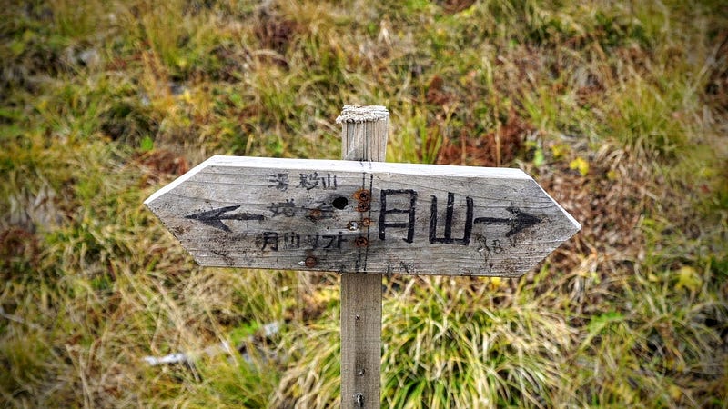 An old and battered wooden sign shows the way to Mt. Yudono, Mt. Ubagatake, and the Gassan Ski Lift to the left, and the summit of Mt. Gassan to the right.