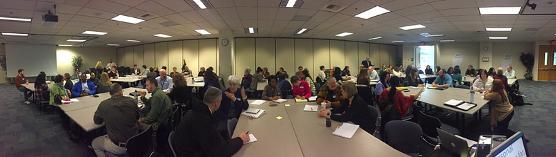 A very large meeting in place, with many people clustered around many tables within a large room. Everyone is sitting.