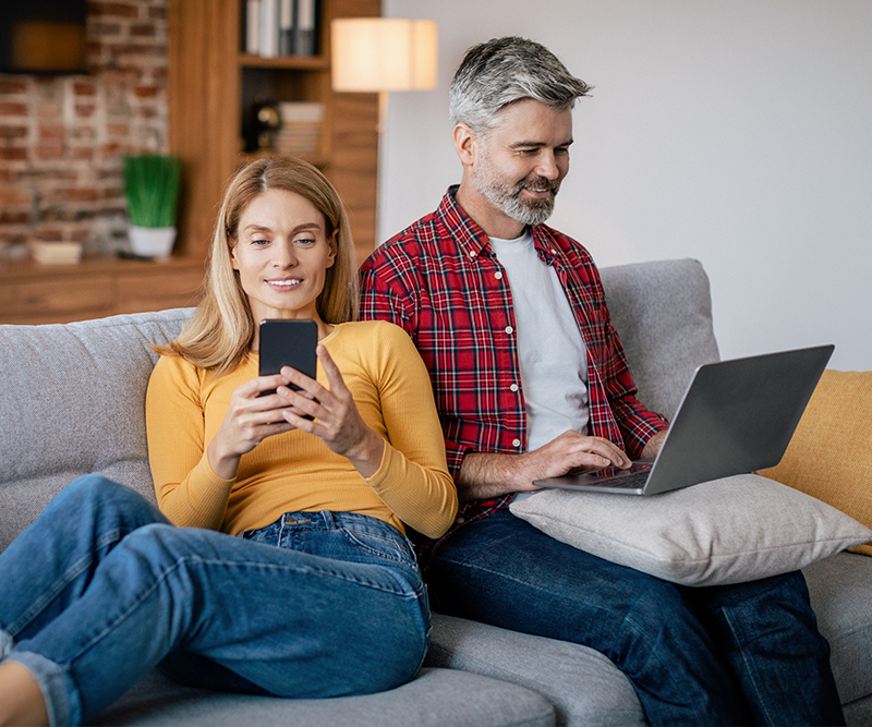 A man and woman using their smartphone and laptop in the comfort of their home with Telsim NBN home Internet.