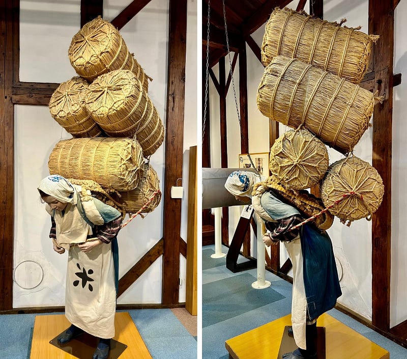 Model of a woman carrying 5 bales of rice on her back.