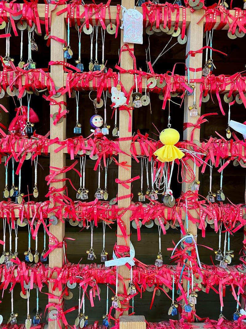 Wooden frame covered in red ribbons holding coins, cubes of glass, and toys, on a shrine on Mount Haguro.